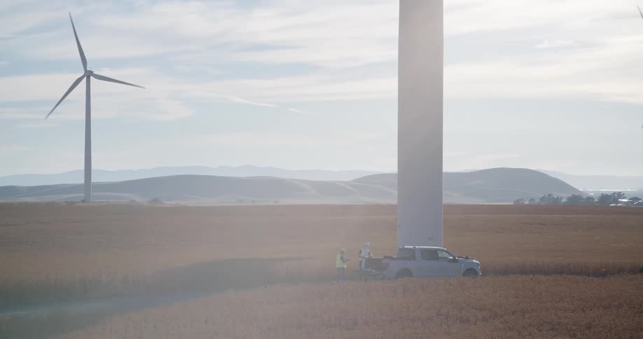 Two engineers in safety gear work from the back of a truck at the base of a wind turbine in a sprawling field