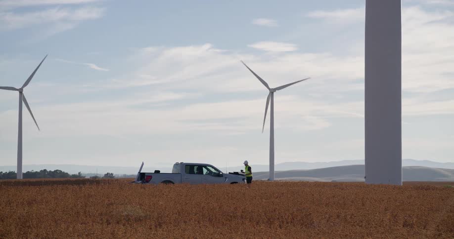 Two engineers in safety gear work from their truck while performing maintenance at a sprawling wind farm in a golden field