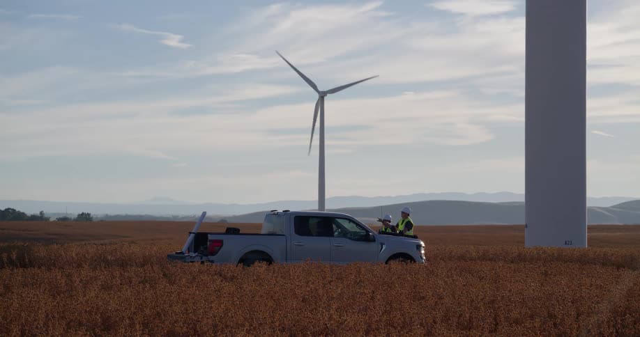 Two engineers in hard hats inspect a wind turbine from their truck in a rural field as the blades spin in the background