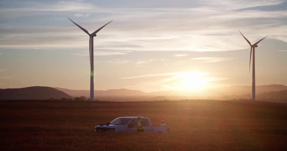 A wide panning shot reveals wind farm engineers with their truck in a vast field of spinning turbines at sunset