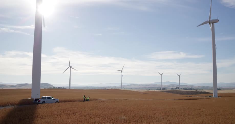 Two engineers inspect a massive wind turbine on a sprawling wind farm in a sunny, golden field