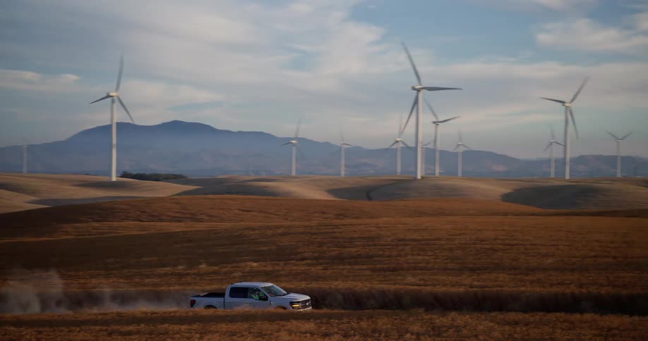 A white pickup truck kicks up dust while driving along a dirt road through rolling golden fields with a wind farm on the horizon