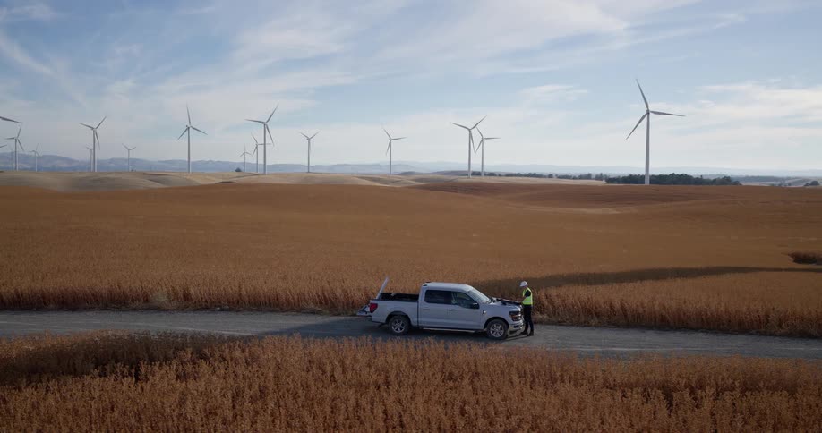 Wide aerial shot pans across a large wind farm where an engineer works from his truck in a vast golden field
