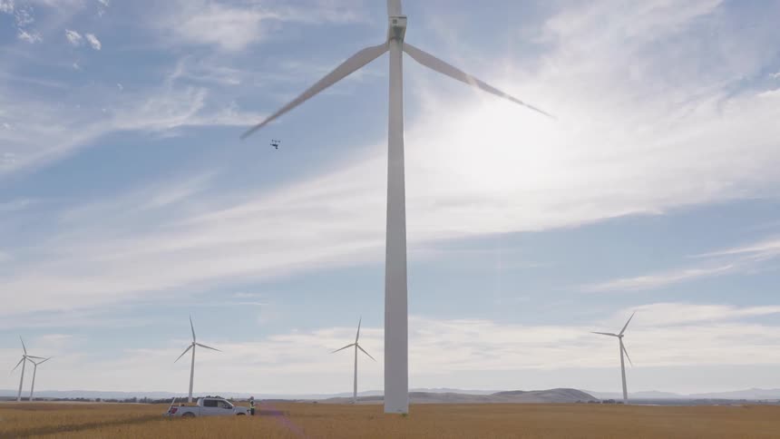 A drone ascends a towering wind turbine for an inspection as engineers watch from below in a sunny, golden field