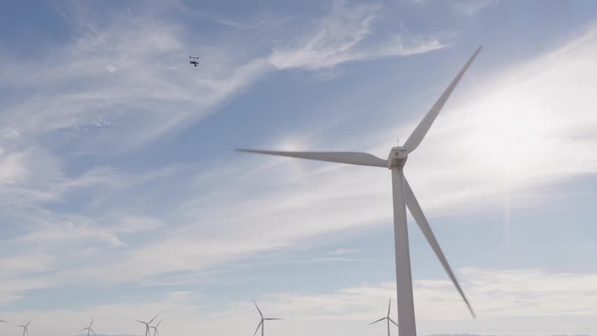 A drone flies away from a large spinning wind turbine at a sunny wind farm