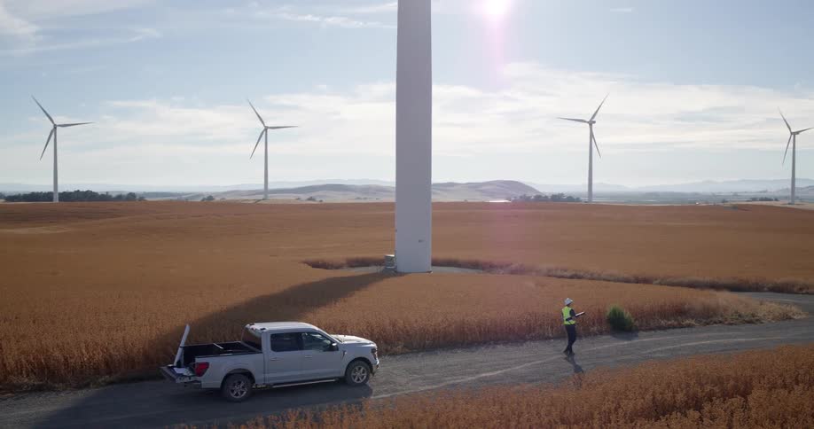 An aerial view shows a wind turbine technician inspecting a wind farm in a vast, golden agricultural field