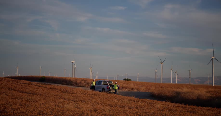 Two wind farm technicians prepare equipment from their truck on a dirt road amidst rolling golden hills at dusk