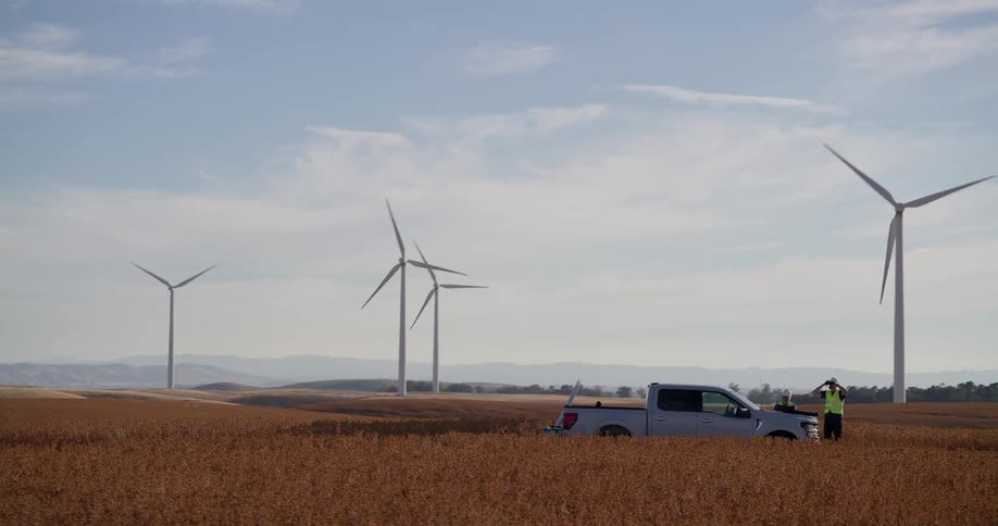 A panning shot reveals two engineers working by their truck amidst a golden field, with a vast wind farm generating clean energy across the rolling hills