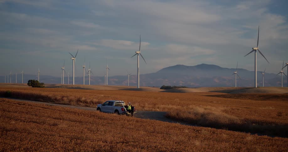 Two wind turbine technicians prepare for work by their truck on a vast wind farm during a golden sunset