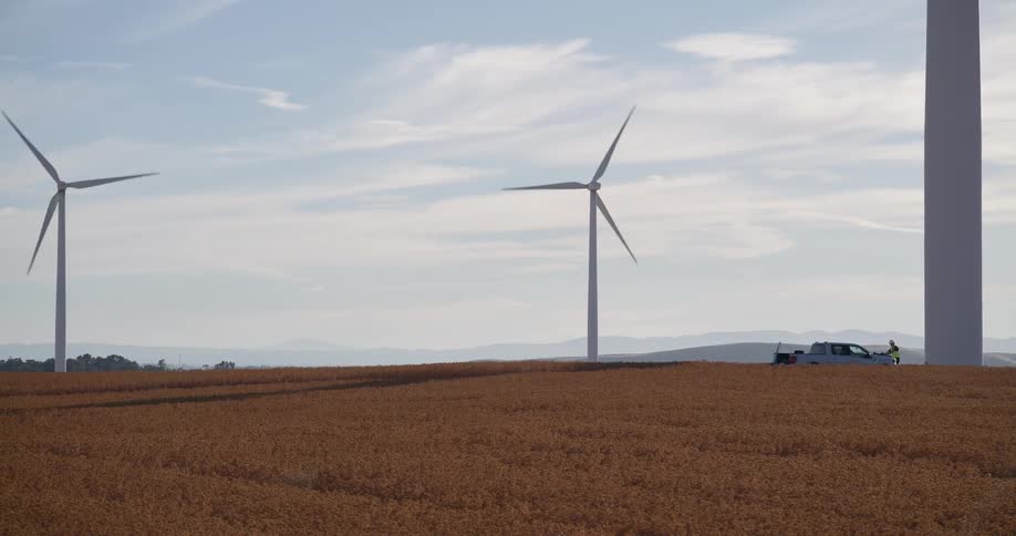A distant shot of a maintenance worker and their truck at the base of a giant wind turbine in a sprawling agricultural field