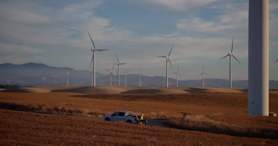 A technician stands by their truck on a dirt road in a field of spinning wind turbines on rolling hills at sunset