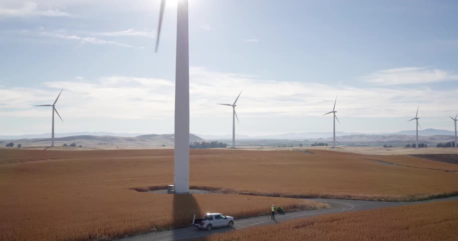 A technician stands by his pickup truck at the base of a massive wind turbine on a sunny day at a wind farm in a golden agricultural field