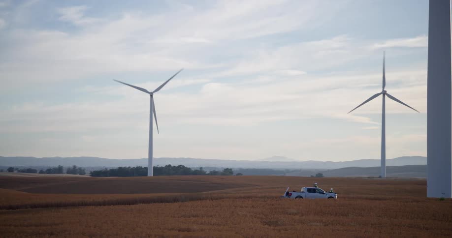 A service truck drives through a golden field on a wind farm with massive turbines spinning against a hazy sky