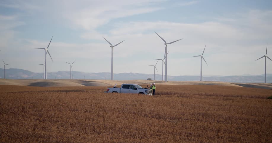 Wide shot of two engineers standing next to their truck in a golden field at a wind farm