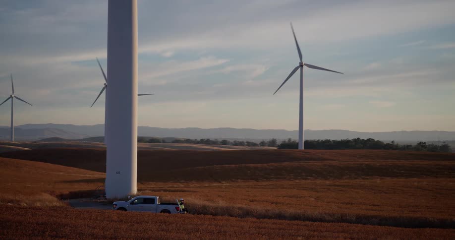 Wide panning shot of a vast wind farm on rolling golden hills with technicians servicing a turbine from a pickup truck