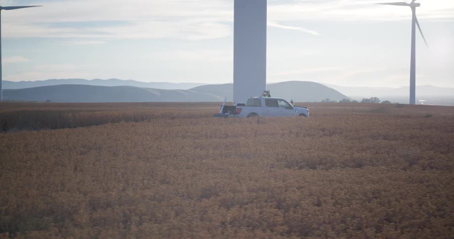 A slow pull back reveals a work truck parked in a vast golden field beneath massive, spinning wind turbines