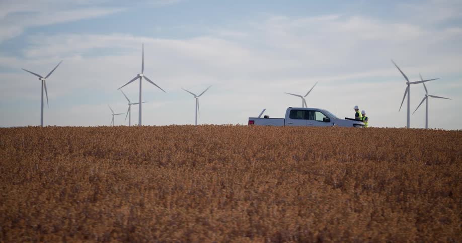 Two wind turbine technicians stand by their truck in a golden field overlooking a vast wind farm