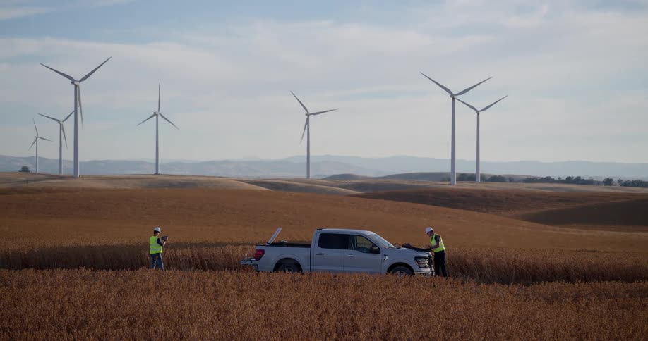 Two wind turbine engineers perform a site inspection in a vast golden field with numerous spinning turbines in the background