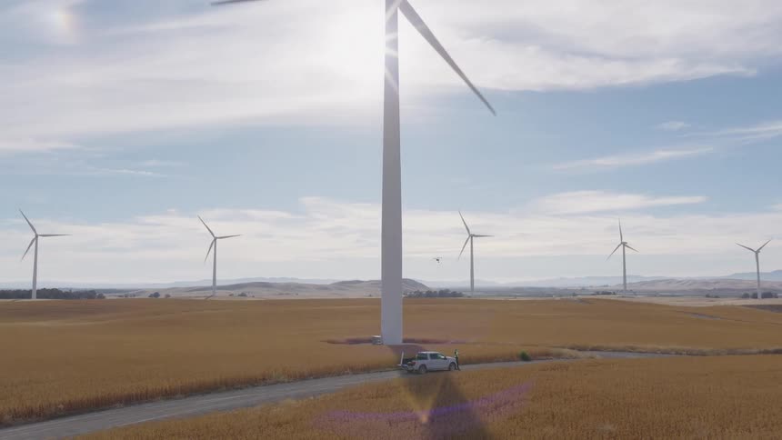 A drone flies over a vast wind farm with spinning turbines in a golden field on a sunny day as a truck is parked on a dirt road