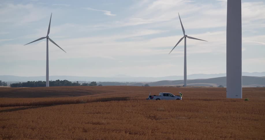 A technician stands beside their truck at the base of a towering wind turbine in a vast golden field as the camera slowly zooms out