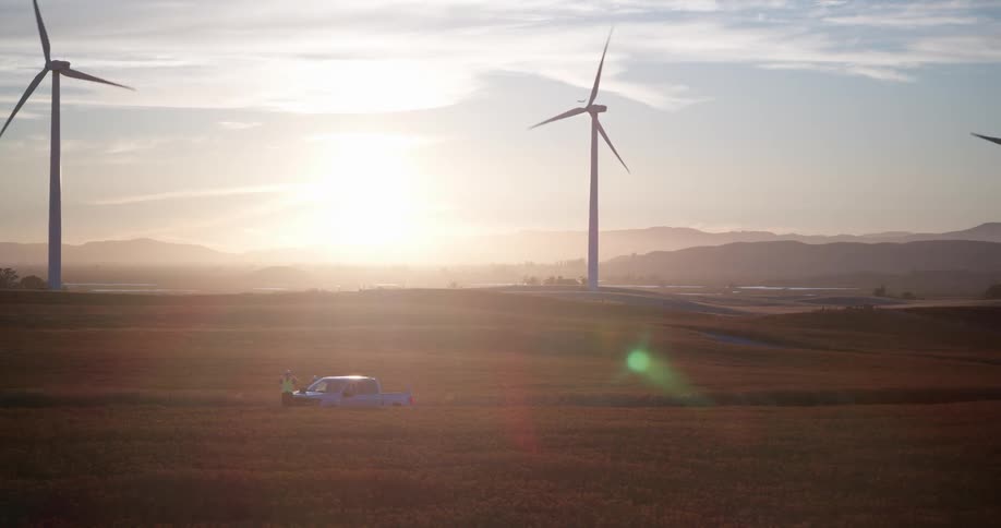 An aerial view captures a technician standing by a truck in a vast field, with large wind turbines spinning against a beautiful sunrise