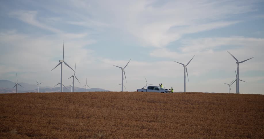 Engineers stand by their truck in a vast golden field, surrounded by spinning wind turbines at a renewable energy farm