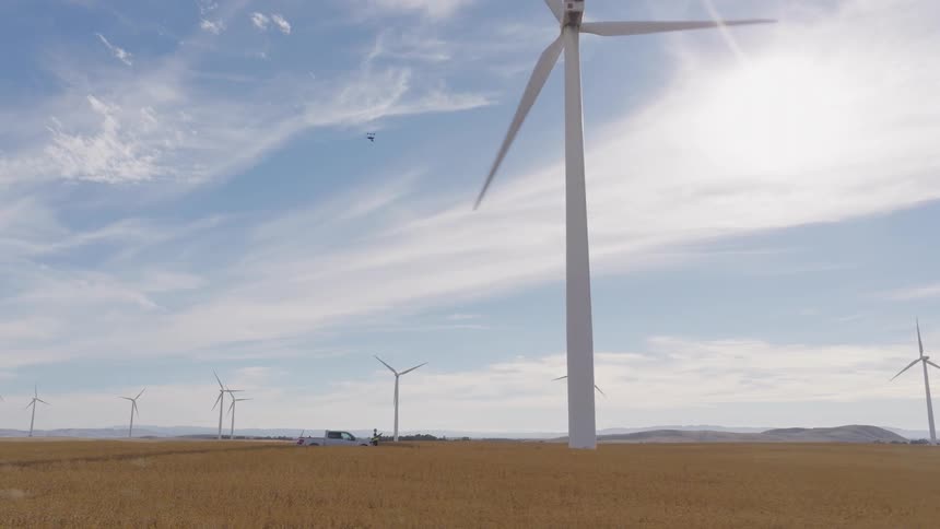 A drone pilot inspects a large spinning wind turbine with a UAV on a sunny day in a golden field full of wind turbines