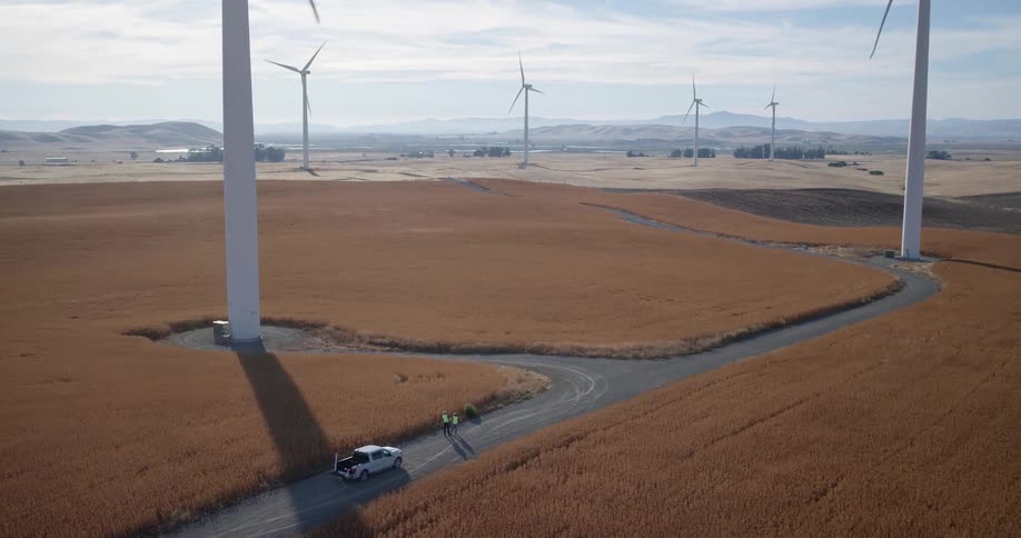Aerial view of two wind turbine technicians standing by their truck in a rural field as the camera pulls back to reveal the expansive wind farm