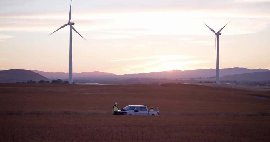 A wind turbine technician stands by a work truck in a vast golden field as windmills spin during a scenic sunset
