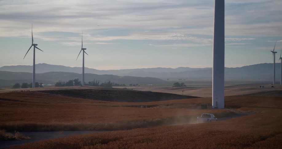 A white pickup truck drives on a dusty road through a field of wind turbines on rolling hills at sunset