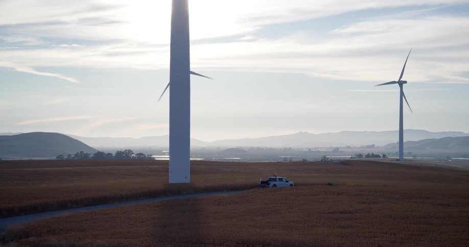 A white truck is parked at the base of a massive wind turbine spinning in a golden field with another turbine visible in the hazy background