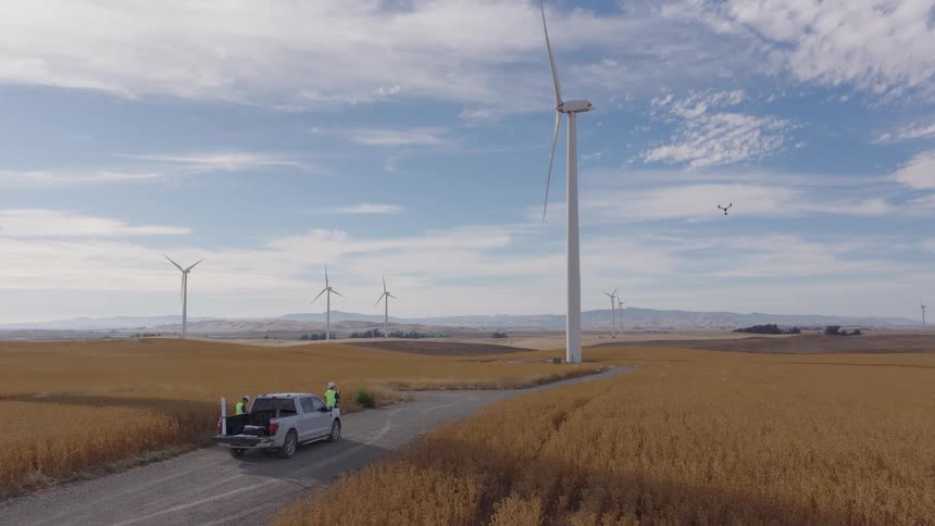 Engineers operate a drone to perform maintenance and inspection on a towering wind turbine at a modern wind farm