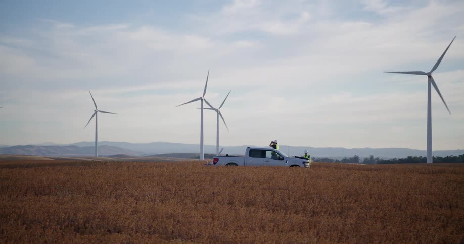 Slow zoom out from a work truck reveals a massive wind farm with technicians on site in a vast golden field