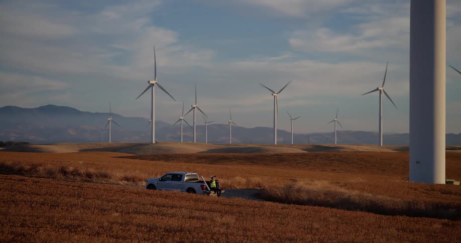 A worker stands by a pickup truck at a vast wind farm with spinning turbines across golden rolling hills
