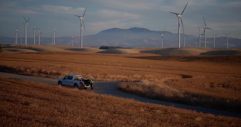 Engineers stand by their truck at a vast wind farm with turbines spinning across golden rolling hills as the sun sets