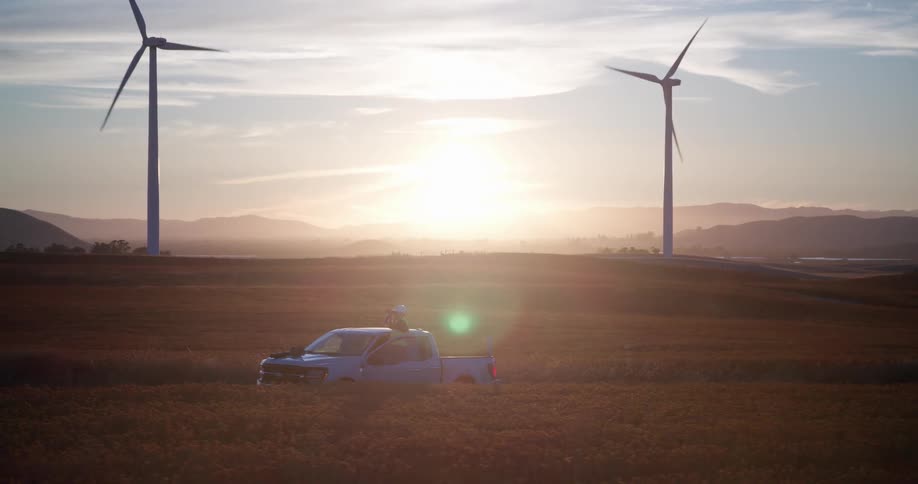 A lone technician stands by their truck at a vast wind farm, with turbines spinning gracefully in a golden field as the sun sets