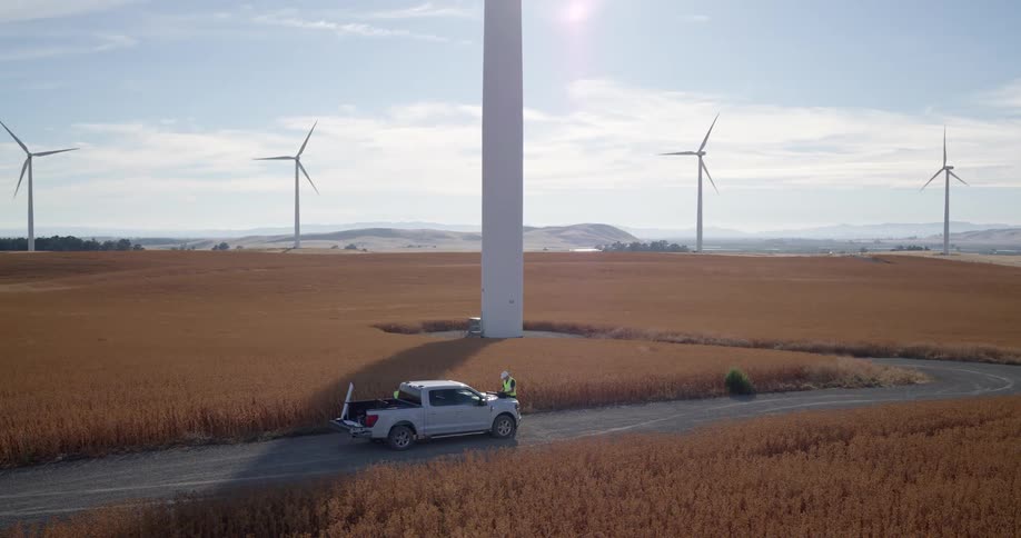 Wide aerial view of two technicians by a truck at the base of a massive wind turbine in a sprawling golden field on a sunny day