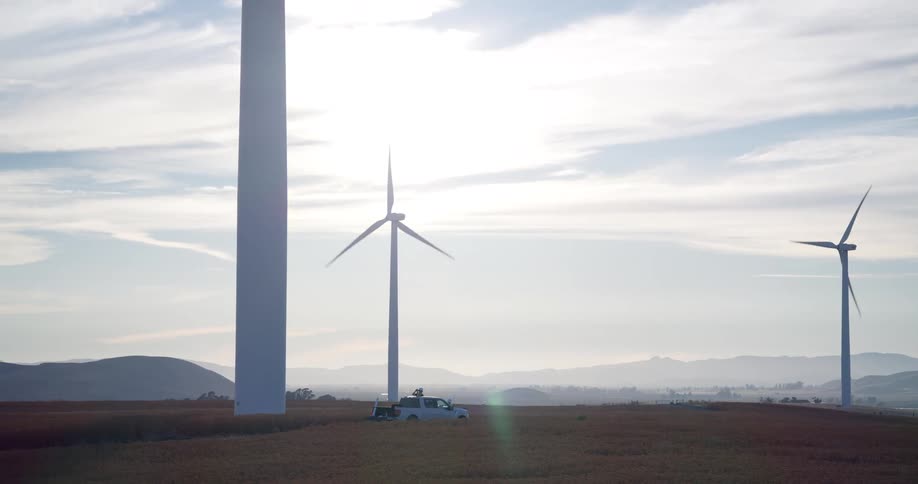 A service truck drives through a field to the base of a towering wind turbine as workers get out to begin maintenance