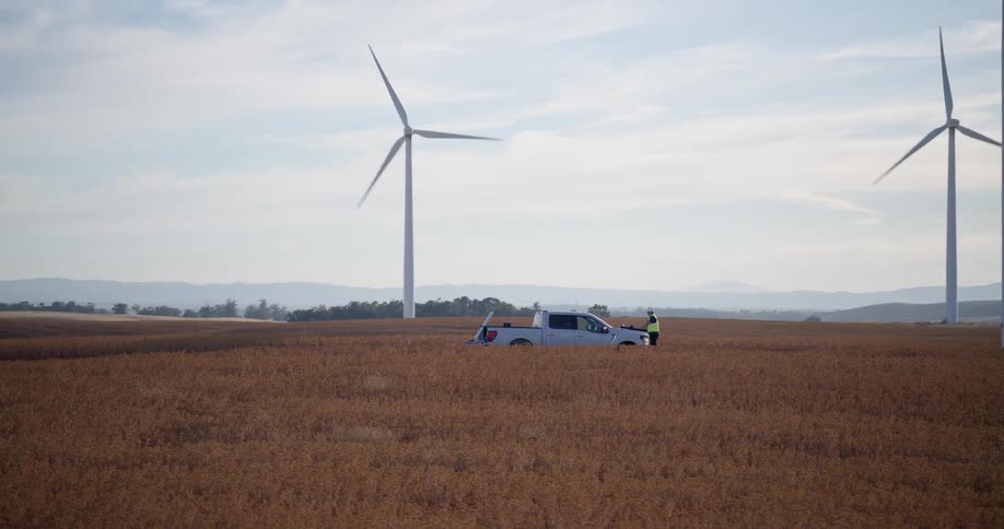 A maintenance worker stands next to a pickup truck at the base of a giant wind turbine in a vast golden field as the camera slowly zooms out