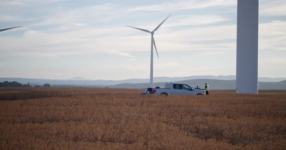 Technicians inspect a giant wind turbine from a truck in a golden field, with a slow pan revealing the expansive wind farm