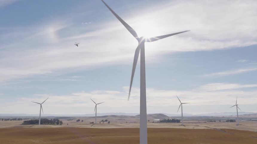 An aerial shot pans up as a drone inspects a spinning wind turbine on a sunny day at a rural wind farm