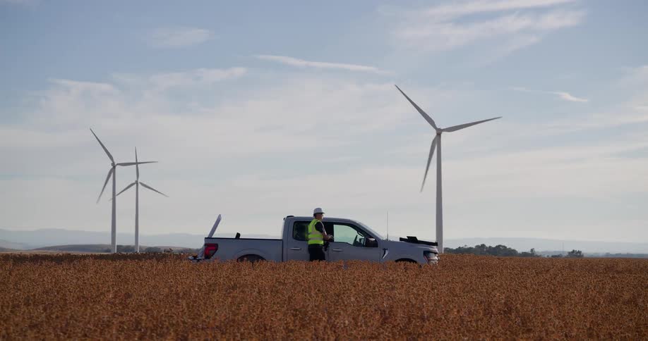 An engineer gets out of his truck in a vast field, inspects the wind farm with binoculars, and sets up equipment on the hood