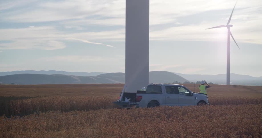 Two engineers in safety vests work at the base of a giant wind turbine in a golden field, with their truck parked nearby and other turbines spinning in the background