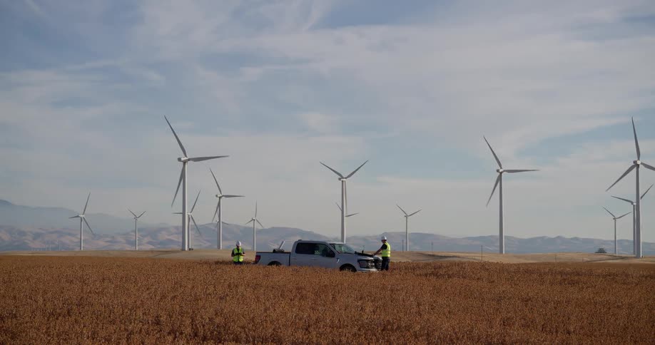 Two engineers collaborate on plans next to their pickup truck amidst a sprawling wind farm in a golden agricultural field