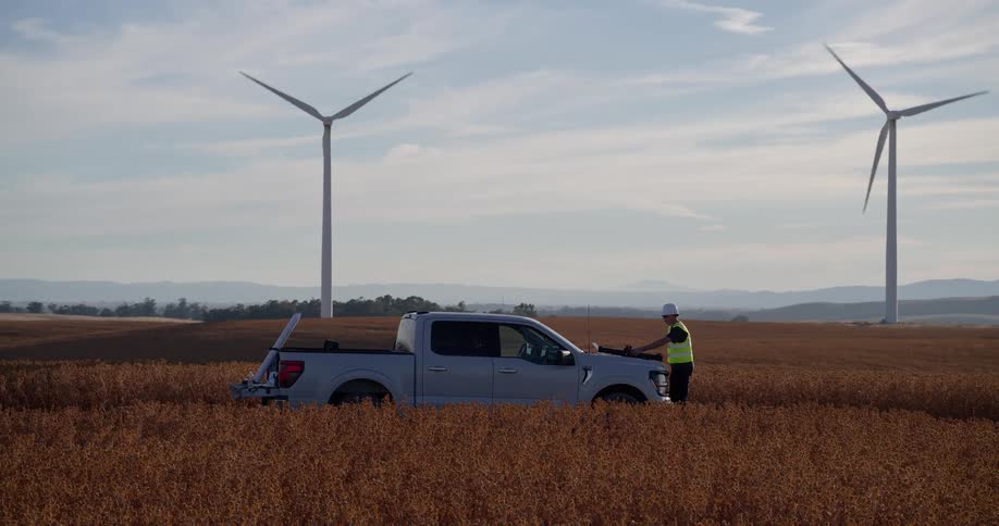 A wind turbine engineer in a hard hat stands by his truck in a golden field, surveying the wind farm on the rolling hills