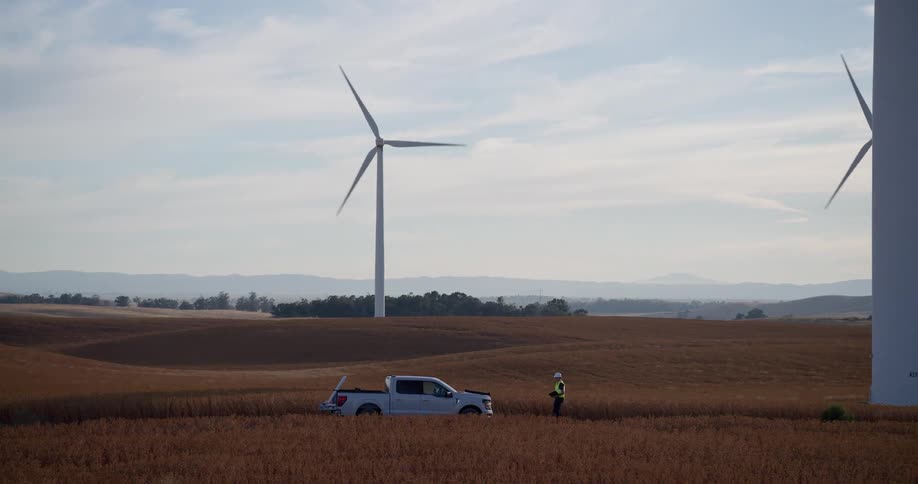 A technician walks from the base of a massive wind turbine through a golden field of crops towards a work truck as the camera pans left across the sprawling wind farm