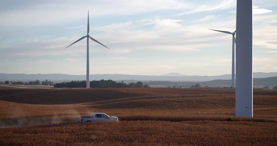 A white pickup truck drives through a golden field on a wind farm, where an engineer gets out to inspect the massive turbines generating renewable energy