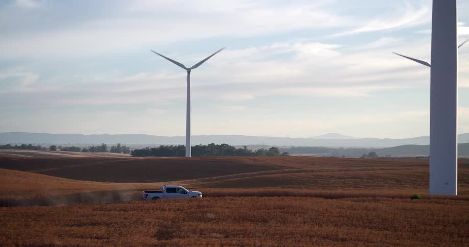 An aerial shot pans to follow a white truck driving through a golden agricultural field on a wind farm