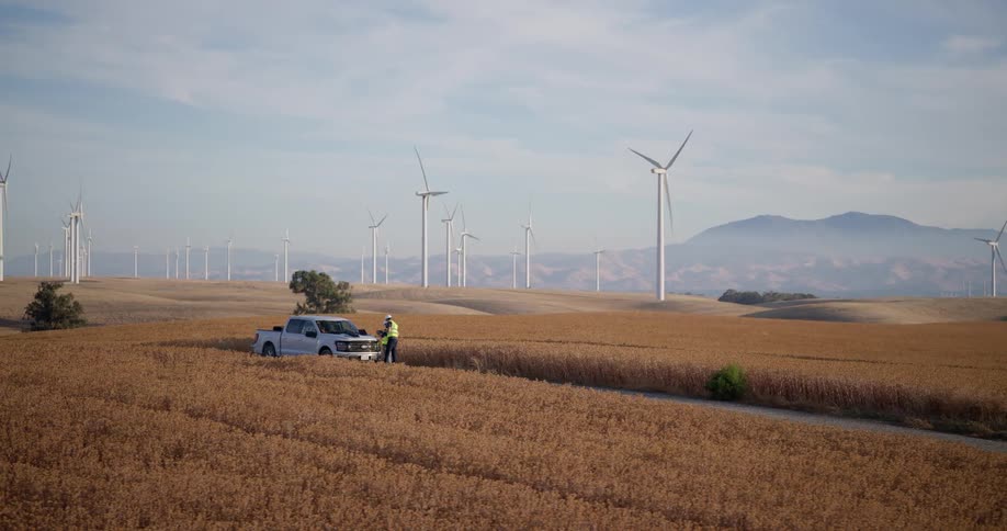 An aerial view pans across an engineer standing next to a truck in a golden field, with a large wind farm generating renewable energy on the rolling hills in the background