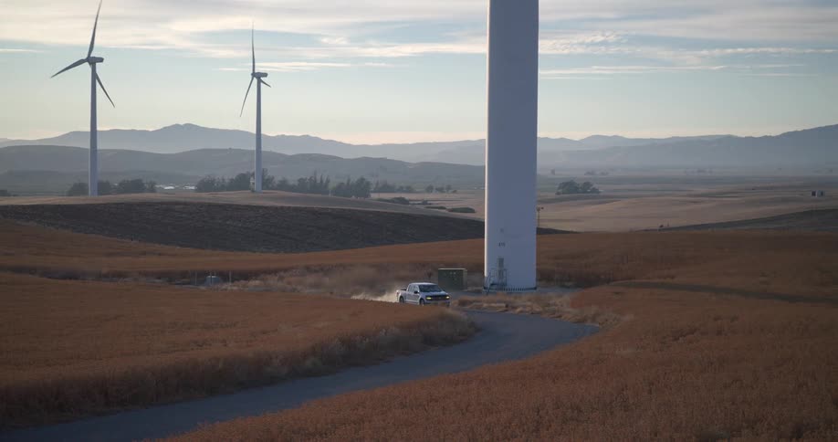 A silver pickup truck drives on a winding dirt road next to spinning wind turbines in a golden field, kicking up dust in slow motion